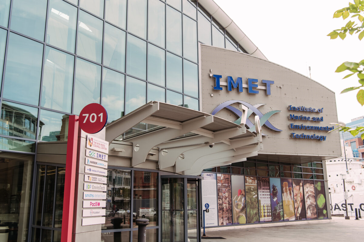 Exterior of a large building; mostly glass windows with a concrete extrusion that says "IMET - Institute of Marine and Environmental Technology" and an overhang above revolving doors.