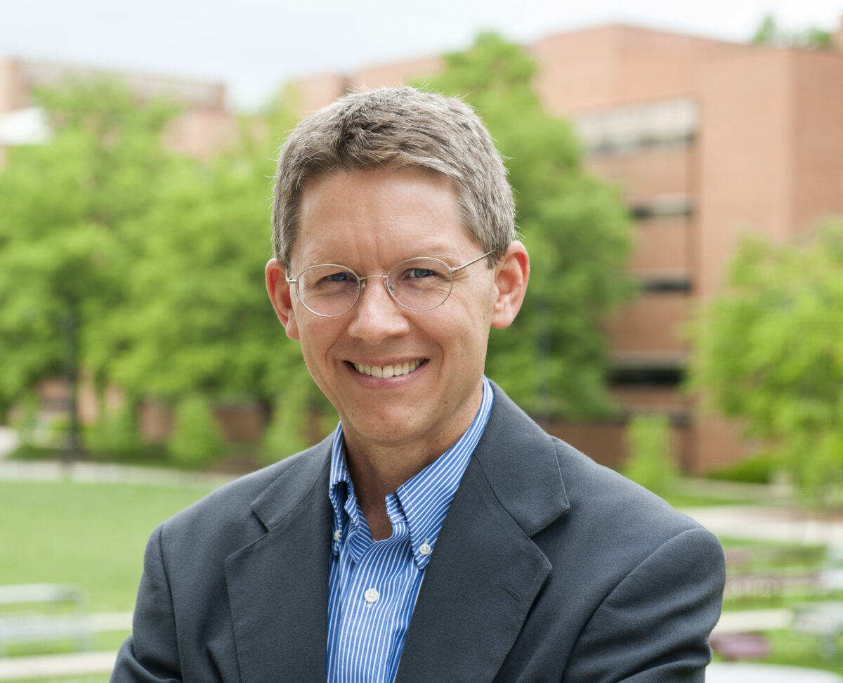 portrait of man standing outdoors in front of green grass, green trees, and red brick building