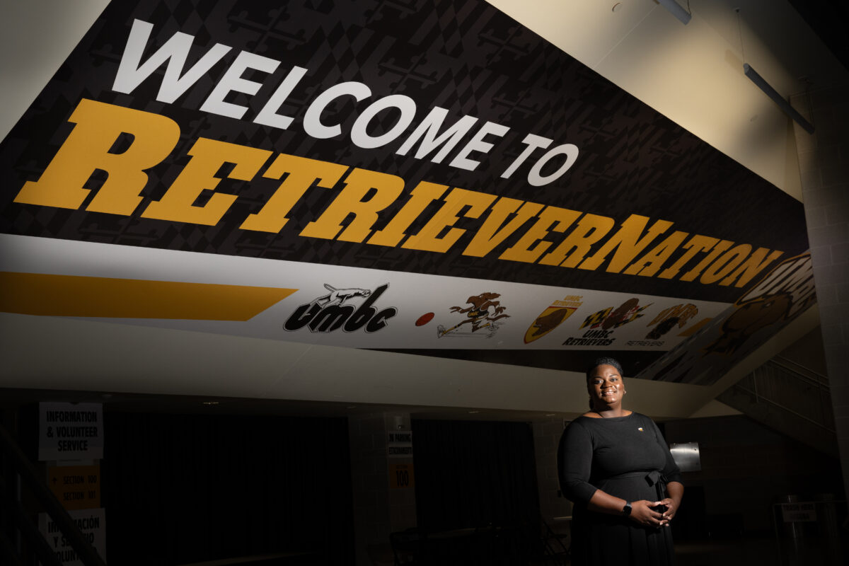 UMBC's AD Tiffany Turner stands in a black dress in front of massive poster spanning slanted ceiling that reads "welcome to retriever nation"