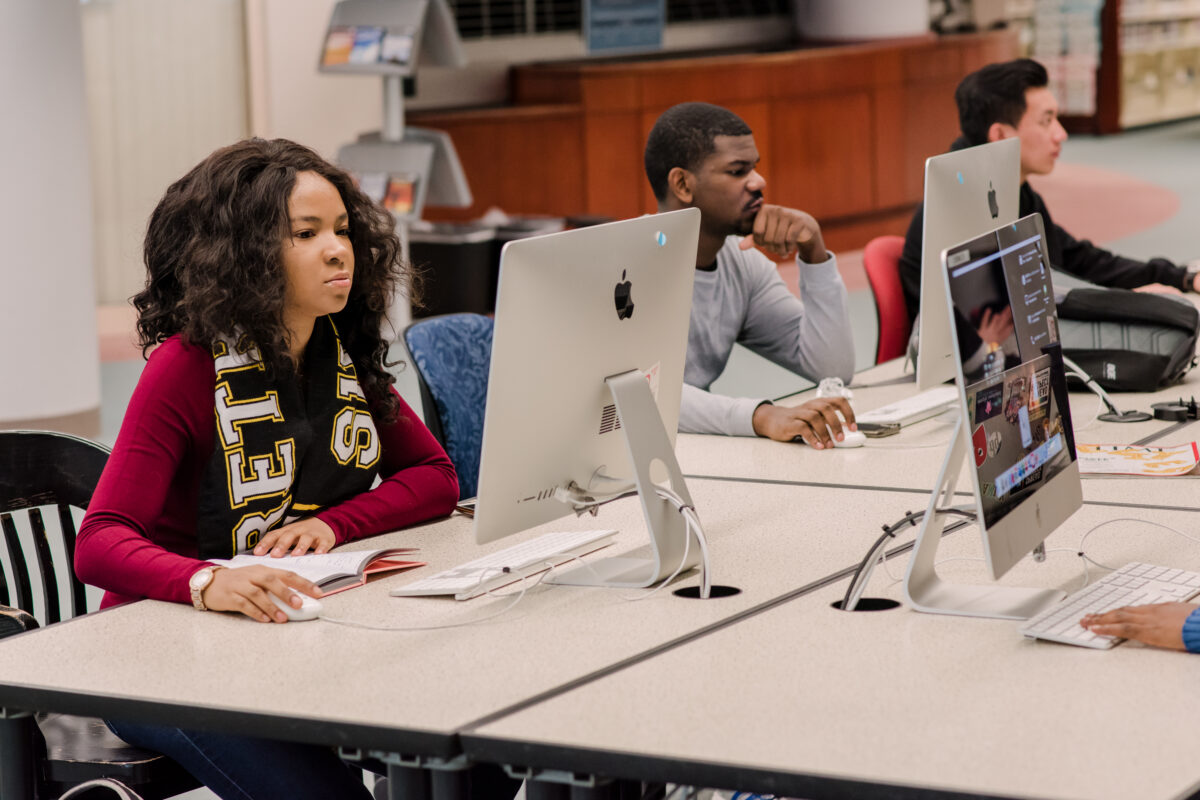 Students sit at table in front of computers. One wears a scarf that says "Retrievers"