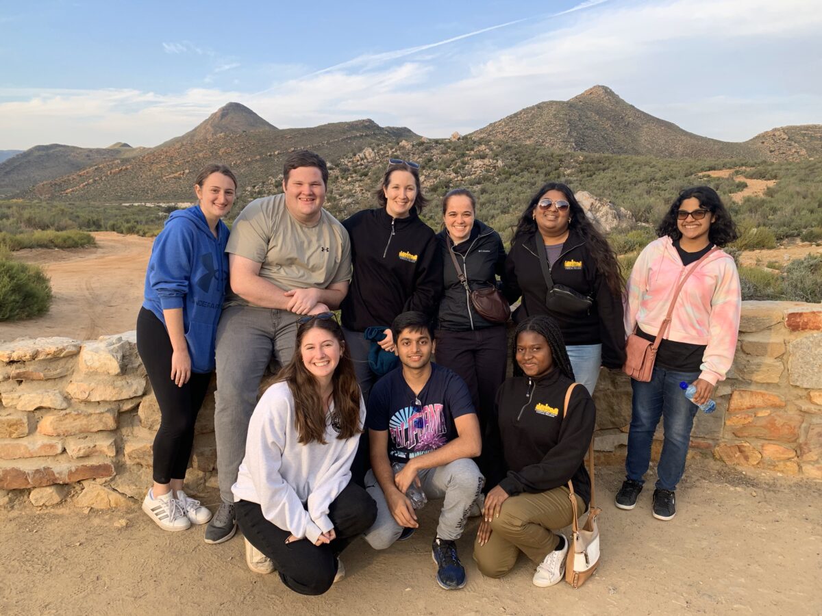 Eight college students stand closely in a group with a professor in front of a stone wall with the South African mountains in the background