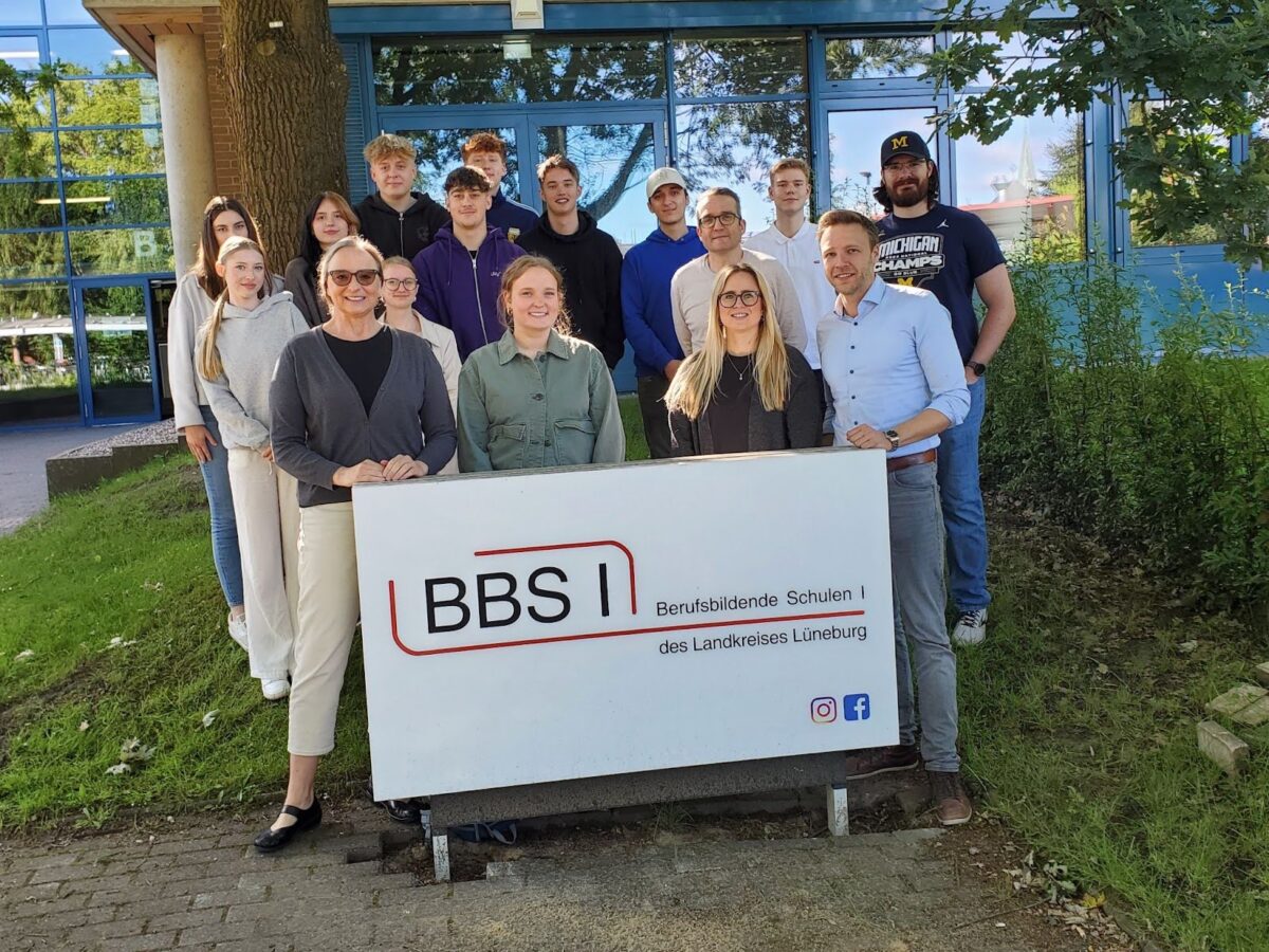 A group of 16 people including teachers and students stand in front of a building holding a banner with German words. Virtual Tandem Conversation Project