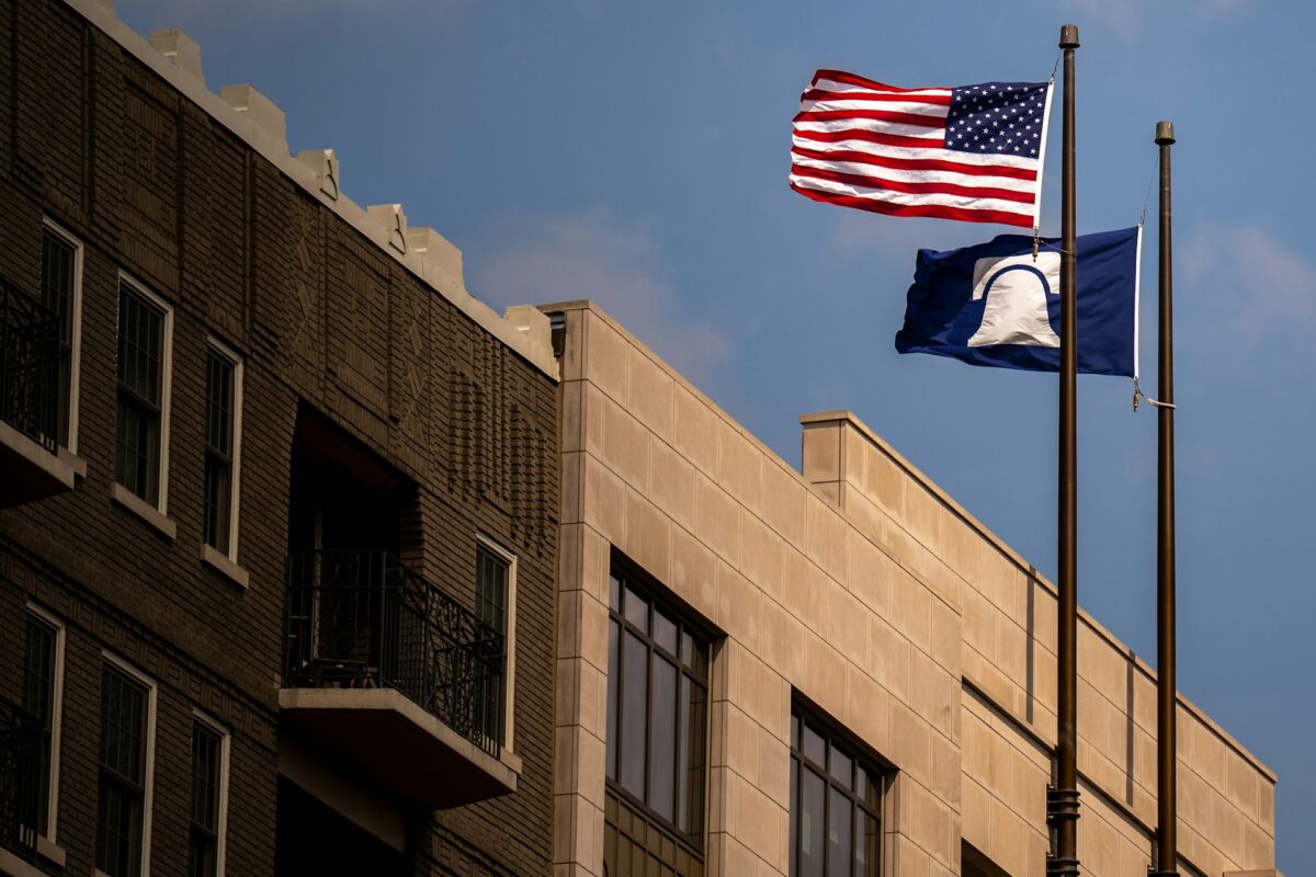 A brick building with barred windows and an upside down American flag and a black flag with a white bell at full mast
