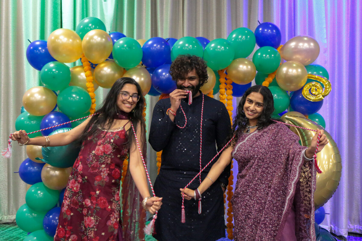 Two women in colorful long dresses and a man in black clothing hold red and white graduation honor cords and stand in front of an arch of red, gold, and green balloons. Retriever Asian Cultural Awards