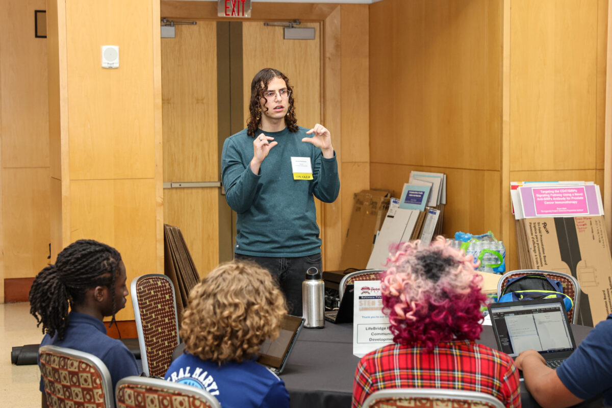 college student, standing and gesturing, speaks to high school students sitting at a table