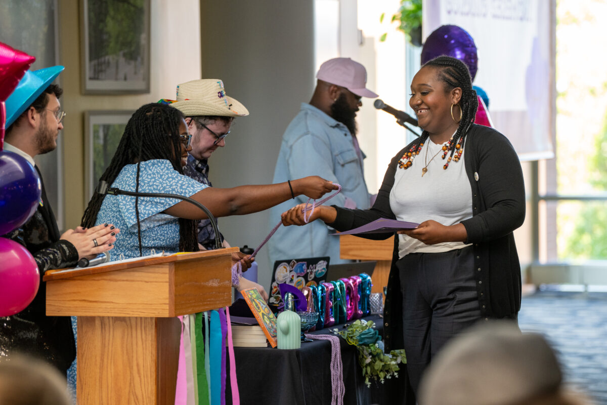 A college student receives an honor cord and recognition certificate from a presenter behind a table decorated with purple and blue bubble letters