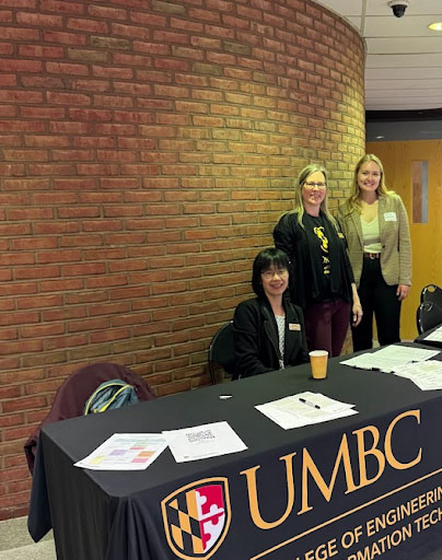 Three people stand near table with UMBC College of Engineering and Information Technology sign