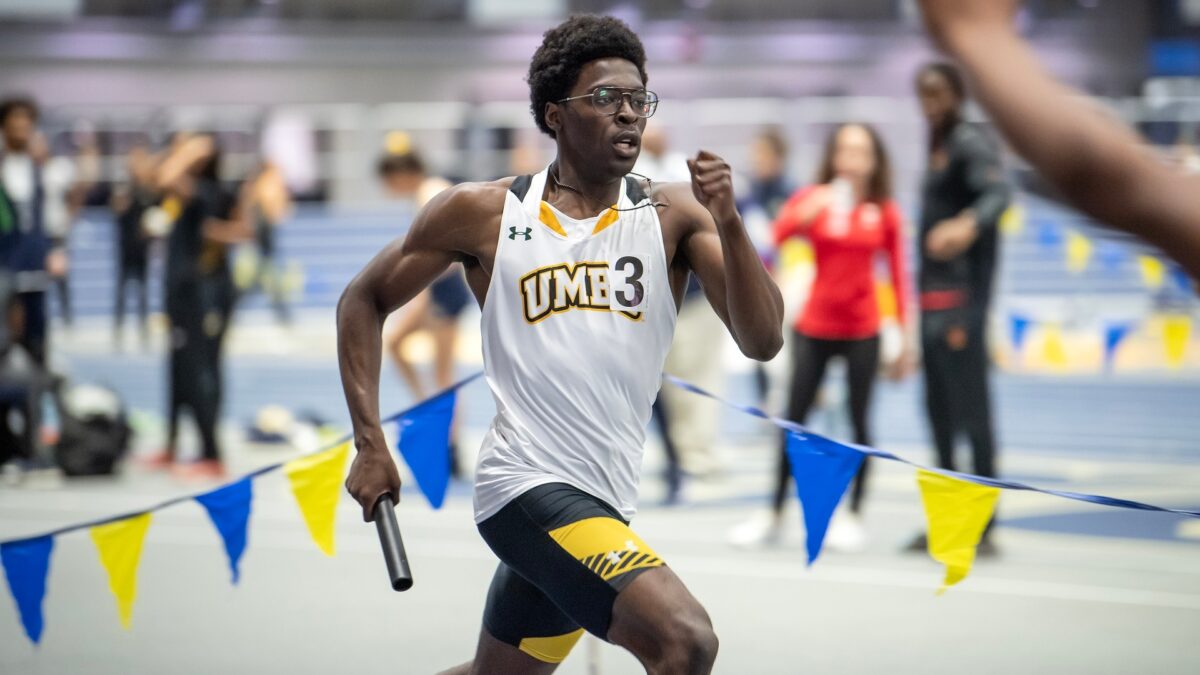 a man in a UMBC jersey runs at a track meet holding a baton